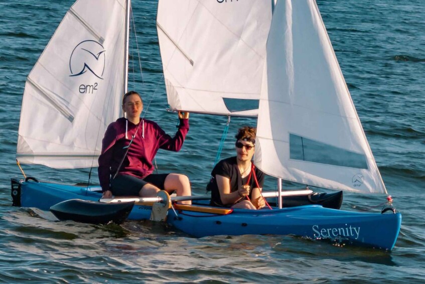 Two people on a sailboat with sails labeled 'em2' and 'Serenity' on a body of water. 1