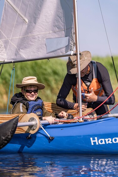 Two people on a sailboat with a blue hull, one wearing a hat and life jacket, and the other adjusting a rope. 5
