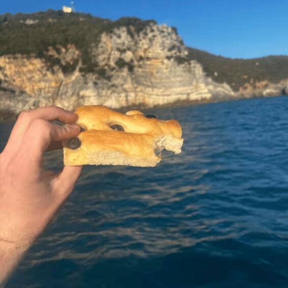 A hand holding a piece of bread with olives, with a coastal cliff and sea in the background. 2