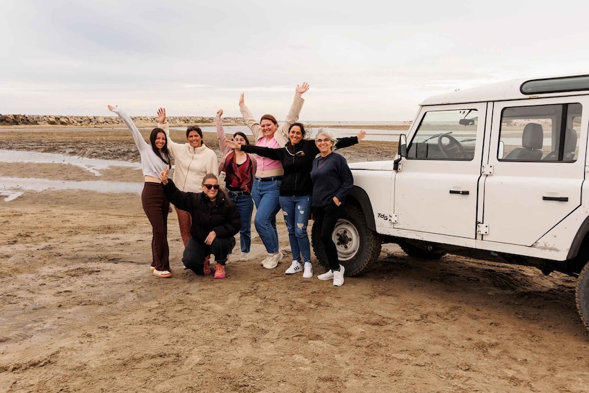 A group of people posing with a white off-road vehicle on a sandy beach. 1