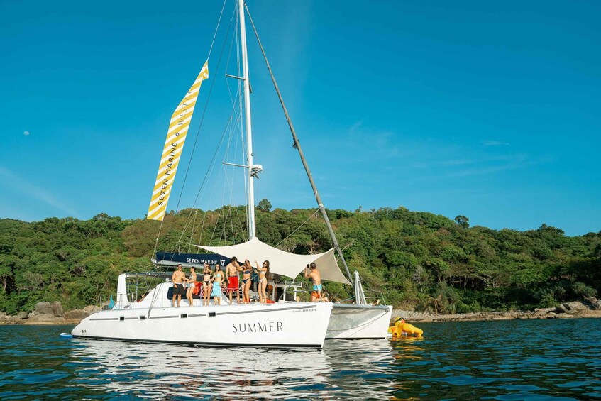 A white yacht named 'SUMMER' with a yellow and white striped sail, docked near a forested shore. 1