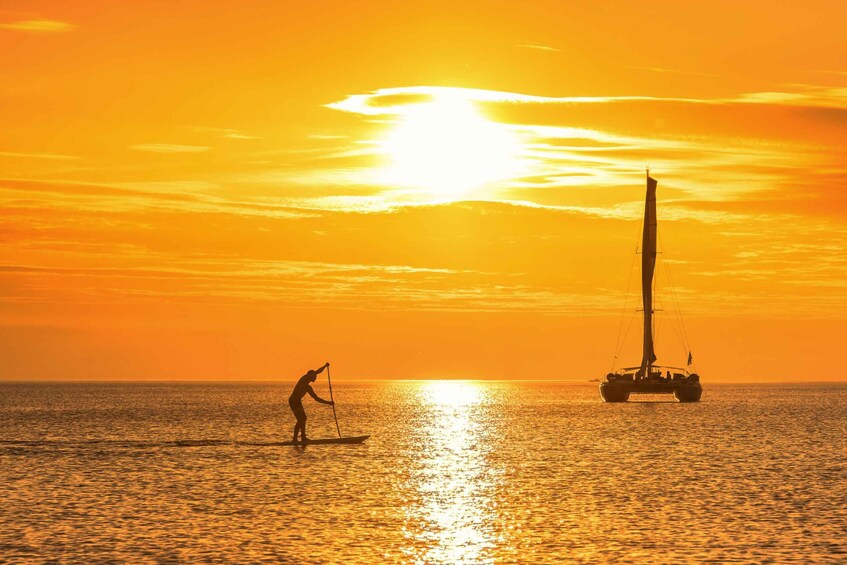 A sunset over the ocean with a sailboat and a person paddleboarding. 5