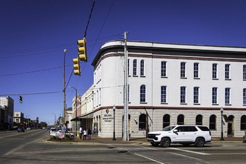 A white building with arched windows and a corner entrance, a white car parked in front, and a traffic light. 5