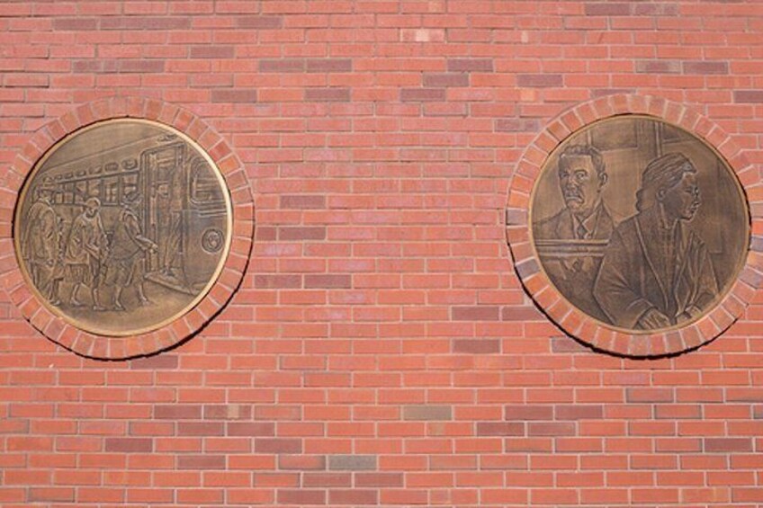 A brick wall with two circular plaques, one depicting a street scene and the other showing two figures. 5