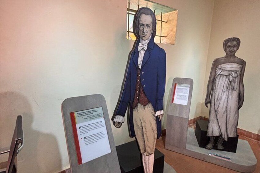 A museum exhibit with two statues, one in a blue coat and the other in a white dress, with informational plaques. 3