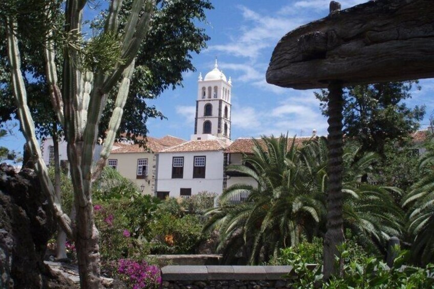 A white building with a bell tower, surrounded by tropical plants and a clear sky. 1