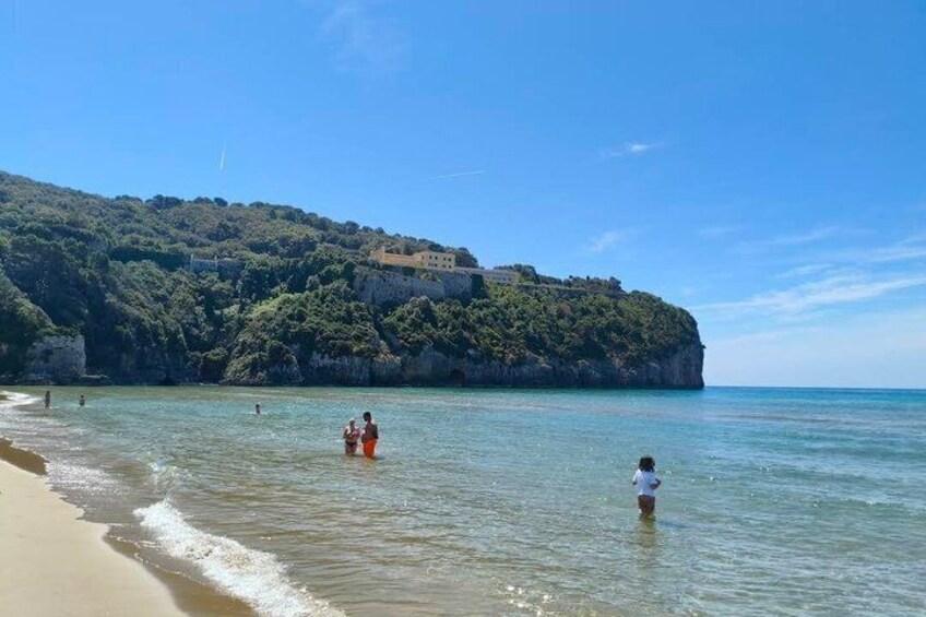 A beach with clear water, people swimming, and a hill with buildings. 4