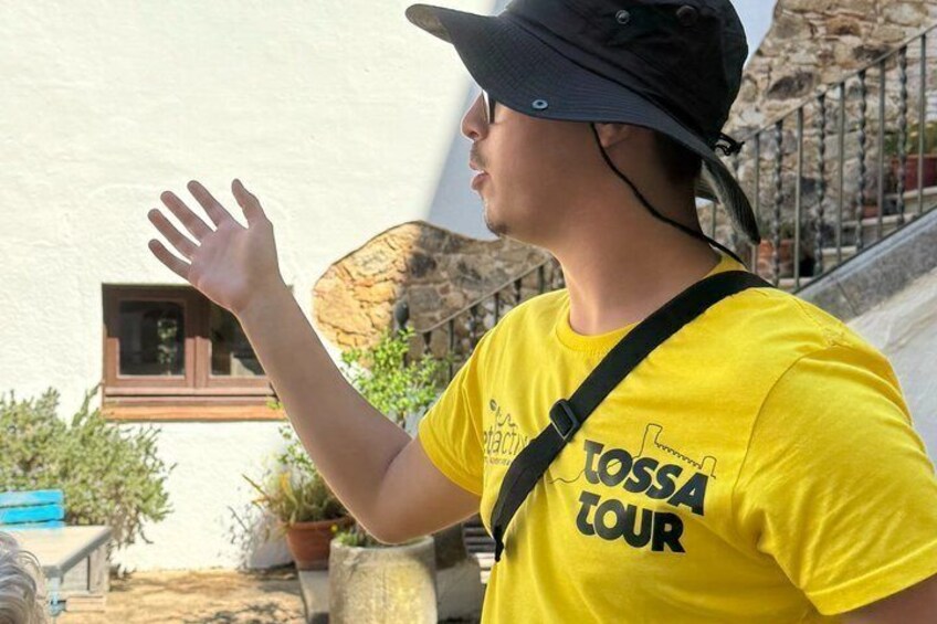 A person in a yellow T-shirt with 'TOSSA TOUR' text, standing outdoors with a stone wall and plants in the background. 5