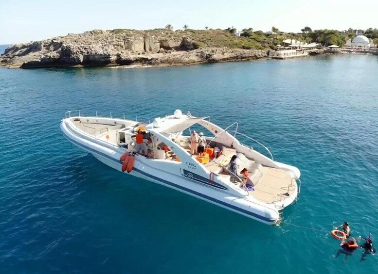 A white motorboat with passengers and life jackets on the water near rocky cliffs and a white building. 1