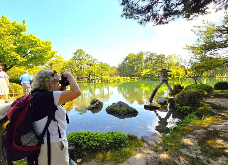 A person taking a photo of a tranquil pond with rocks and a pagoda in a lush garden. 1
