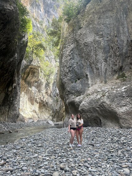 Two people standing on a rocky riverbed with cliffs in the background. 2