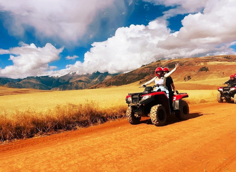 Two people on red ATVs on a dirt road with a mountainous landscape in the background. 1