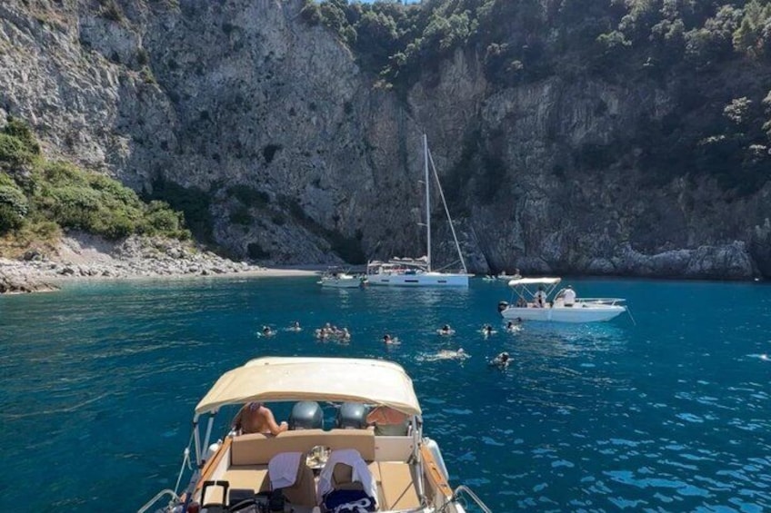 A boat with people swimming in the water, a sailboat, and rocky cliffs in the background. 4