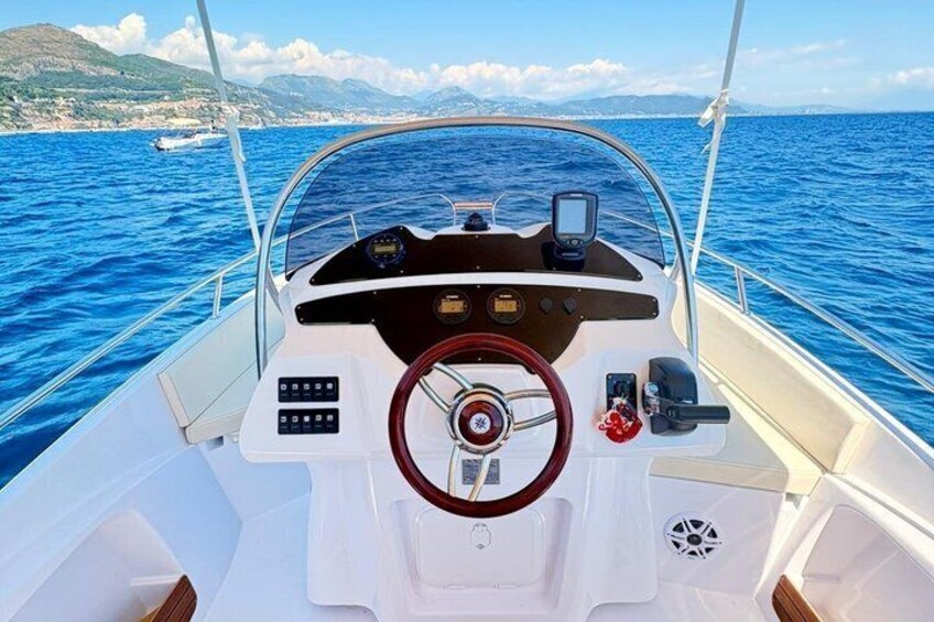 A boat's cockpit with a steering wheel, control panel, and a view of the sea and distant coastline. 2