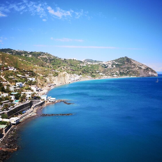 A coastal town with buildings along the shoreline, a clear blue sea, and a hilly landscape in the background. 3