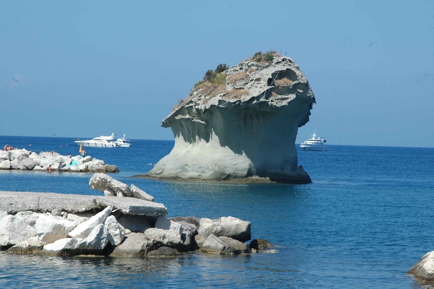 A large rock formation in the sea with boats in the background. 1