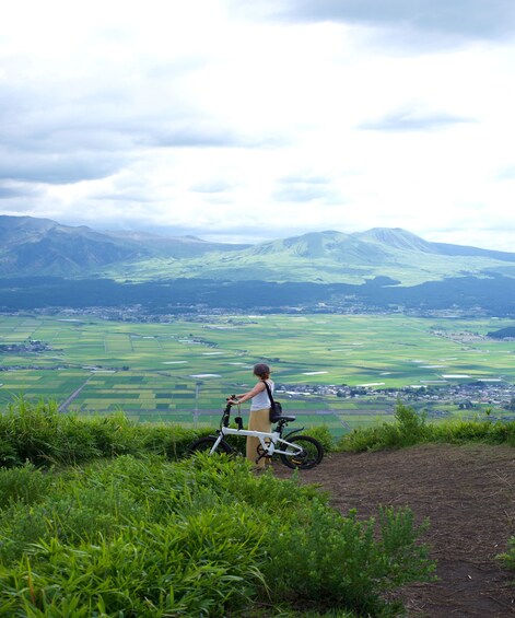 A person standing on a hill with a bicycle, overlooking a valley with fields and distant mountains. 1
