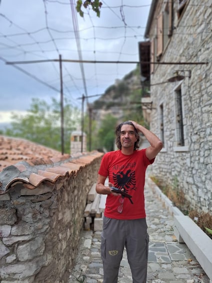 A person standing on a stone-paved path beside a stone wall with a tiled roof, in a narrow alley with buildings and vegetation in the background. 3