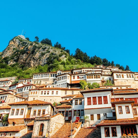 A hillside village with terracotta-roofed buildings, a prominent rocky peak in the background, and a clear blue sky. 1