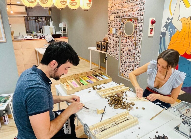 Two people working at a table with wood shavings and tools, surrounded by a wall of photos and a ramen restaurant sign. 5