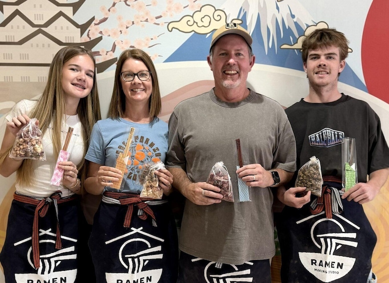Four people holding food items, wearing aprons with 'Ramen' logos, in front of a mural. 4