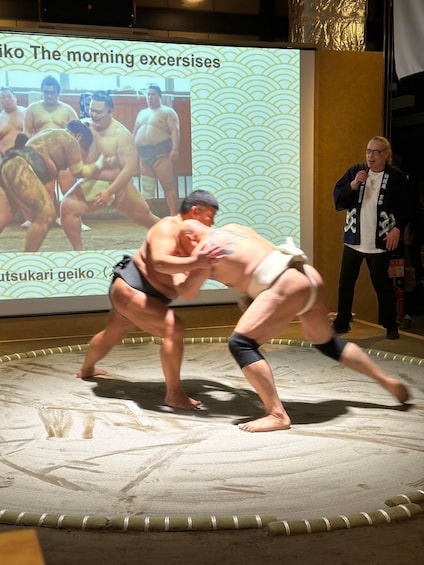 Two sumo wrestlers in a match on a tatami mat with a projection screen in the background. 1