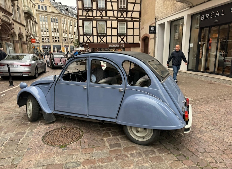 A vintage blue car parked on a cobblestone street with a man walking by. 1