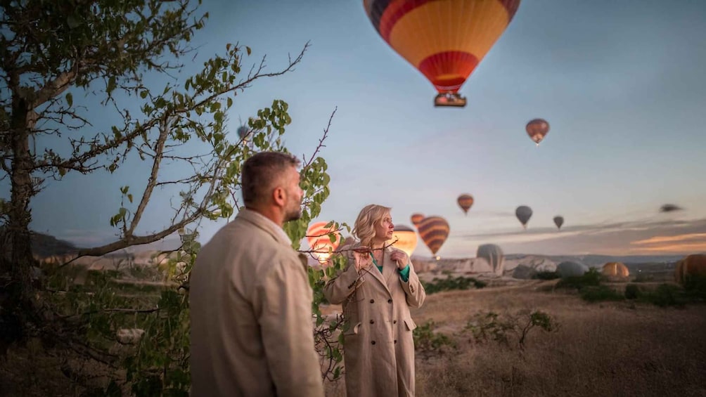 A man and a woman looking at hot air balloons in a rural landscape with trees and open fields. 2