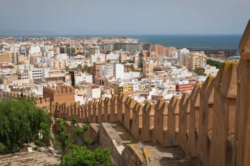 A coastal cityscape with a prominent fortification wall, dense urban buildings, and a clear view of the sea. 2