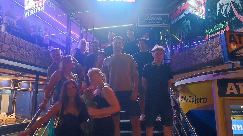 Un gruppo di persone su un nastro trasportatore in un bar con insegne al neon e un bancomat. 2