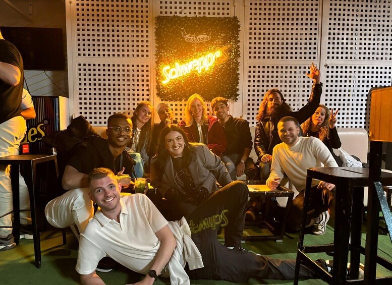 A group of people in a room with a neon sign reading 'Schweppes' on the wall. 2