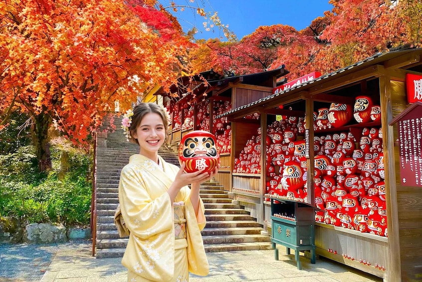 A person holding a traditional mask, standing in front of a shop with red patterned walls and a wooden staircase. 1