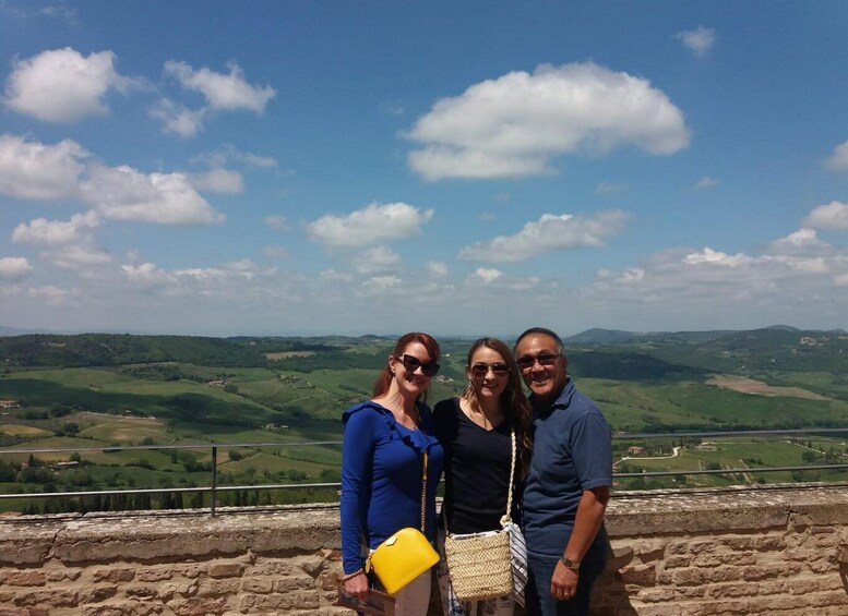 Three people standing on a stone wall overlooking a green valley with scattered hills under a blue sky with clouds. 3