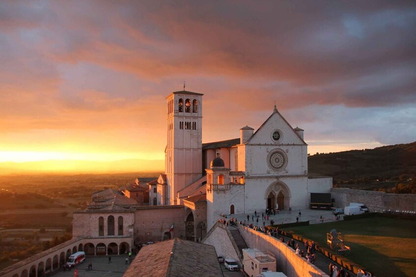 A historic church with a bell tower and a large circular window, surrounded by a fortified wall and a crowd of people. 1