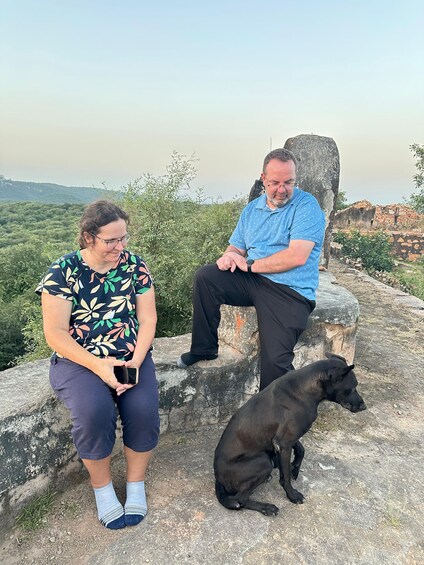 Dos personas sentadas sobre una repisa de piedra, con un perro frente a ellas, contemplando un paisaje montañoso. 5