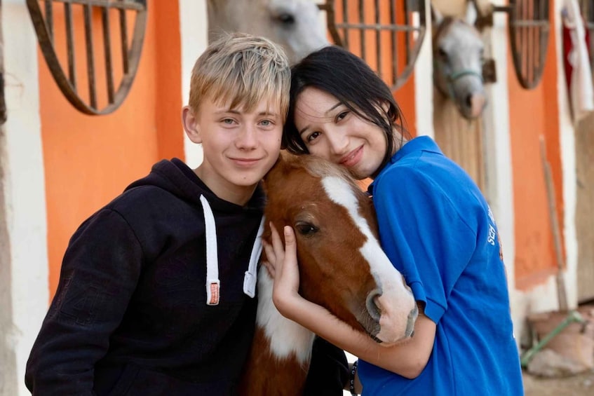 Two people hugging a horse in front of an orange building with metal gates. 4