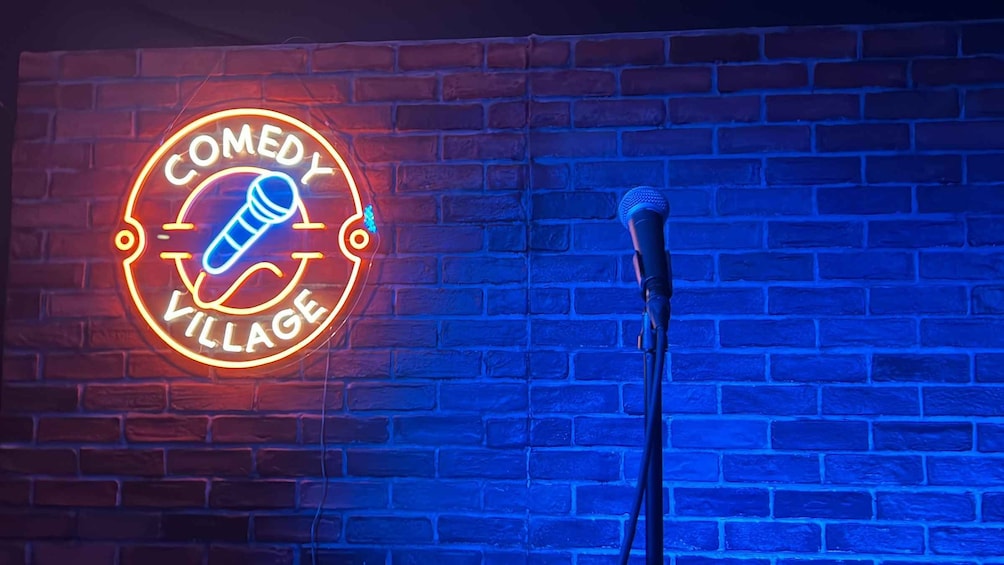 Ein Neon-Schild mit einem Mikrofon und dem Schriftzug 'COMEDY VILLAGE' vor einer Ziegelwand. 3