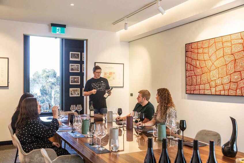 A group of people seated around a table with wine glasses, a man presenting with a bottle and a sign, and a large abstract painting on the wall. 3