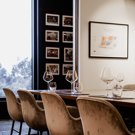 A dining table with empty wine glasses and chairs, a framed certificate on the wall, and a window with a view of trees. 5