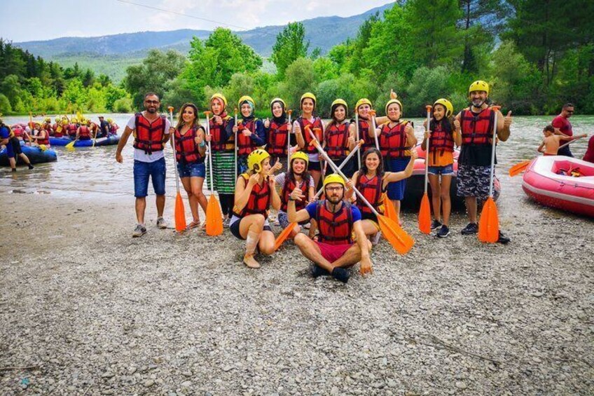 A group of people in life jackets and helmets on a gravel riverbank, with rafts and paddles in the background. 3