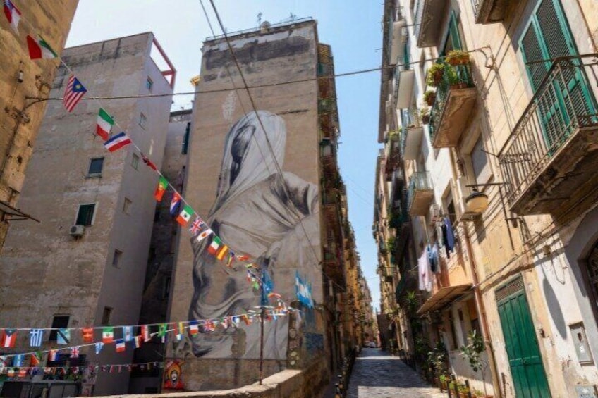 A narrow street with buildings, a mural, and flags. 3