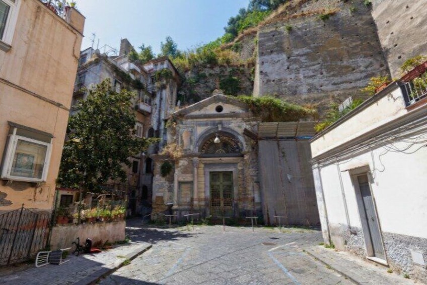 A narrow cobblestone street flanked by historic buildings, a stone wall, and a small church with a bell tower. 2