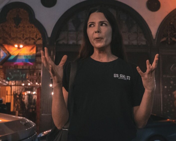 A person in a black t-shirt with 'US GH: 57' text, standing in a dimly lit room with arched ceilings and a car in the background. 3