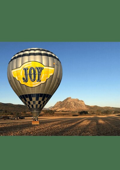 A hot air balloon with 'JOY' written on it, parked on a dirt road with a mountainous landscape in the background. 4