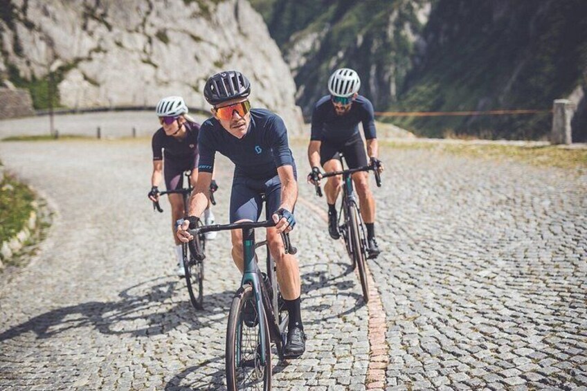 Three cyclists on a cobblestone road with mountains in the background. 3