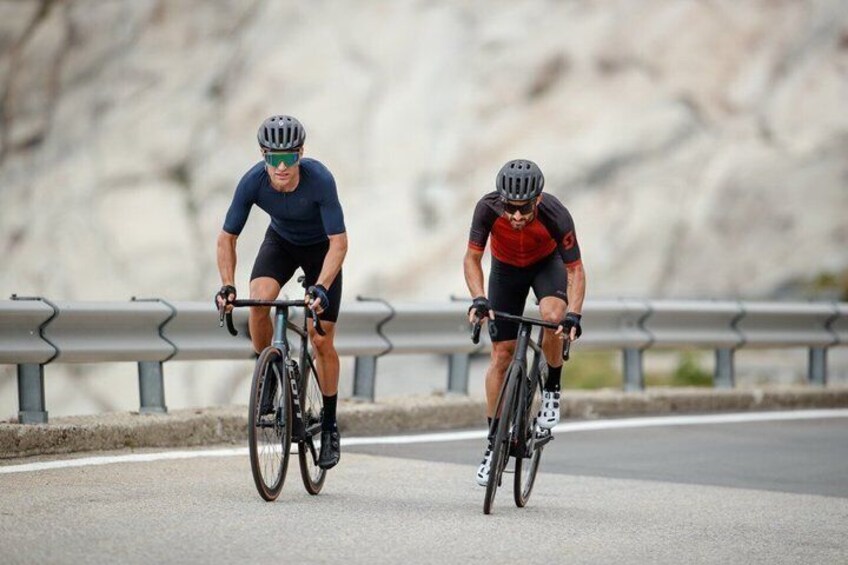 Two cyclists on a paved road with guardrails, wearing helmets and cycling gear. 4