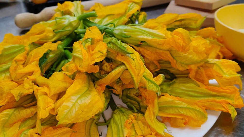 A pile of yellow flowers with green leaves, possibly a type of squash, on a white surface. 1