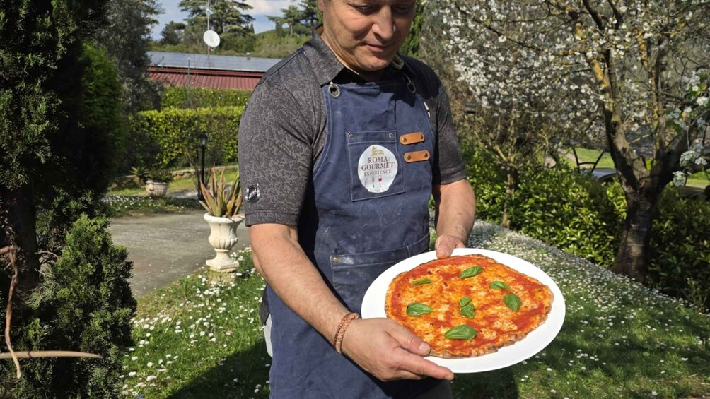 A person holding a pizza with basil leaves, wearing a blue apron with a logo, standing in a garden with trees and a building in the background. 2