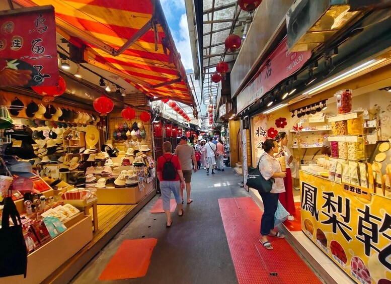 A bustling indoor market with various stalls selling hats and food items, under a red and yellow striped awning. 2