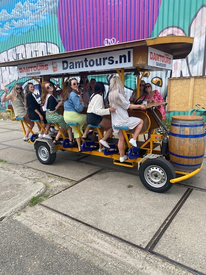 A group of people riding a wooden cart with a barrel attached, labeled 'Damtours.nl,' in front of a colorful mural. 1